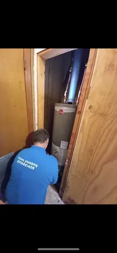 A plumber in a blue shirt works on a water heater in a tight, wooden enclosure.