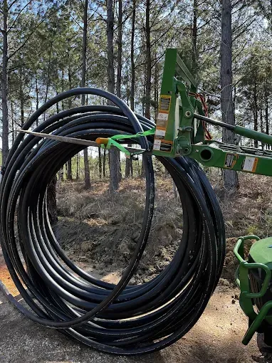 Large coil of black tubing lifted by a green tractor-mounted fork in a wooded area.