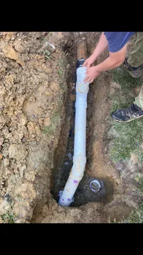 Person installing PVC drain pipe in an outdoor trench; dirt, grass, and purple fittings visible.