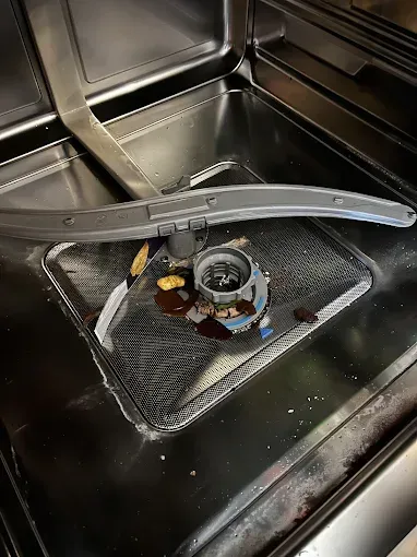 Inside a dishwasher, debris and dark liquid surround the filter and a spray arm.