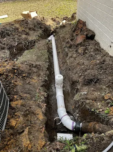 White PVC drain pipe in an open trench, next to a house, amidst dirt and grass.