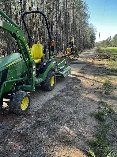 Green tractor on a dirt road; a small backhoe is visible further down the road. Trees line the roadside.