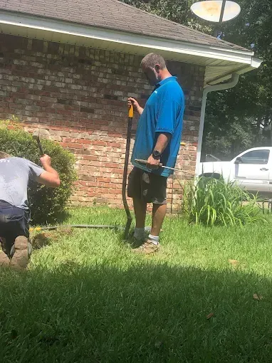 Two men digging in a grassy yard near a brick house. One holds a shovel.