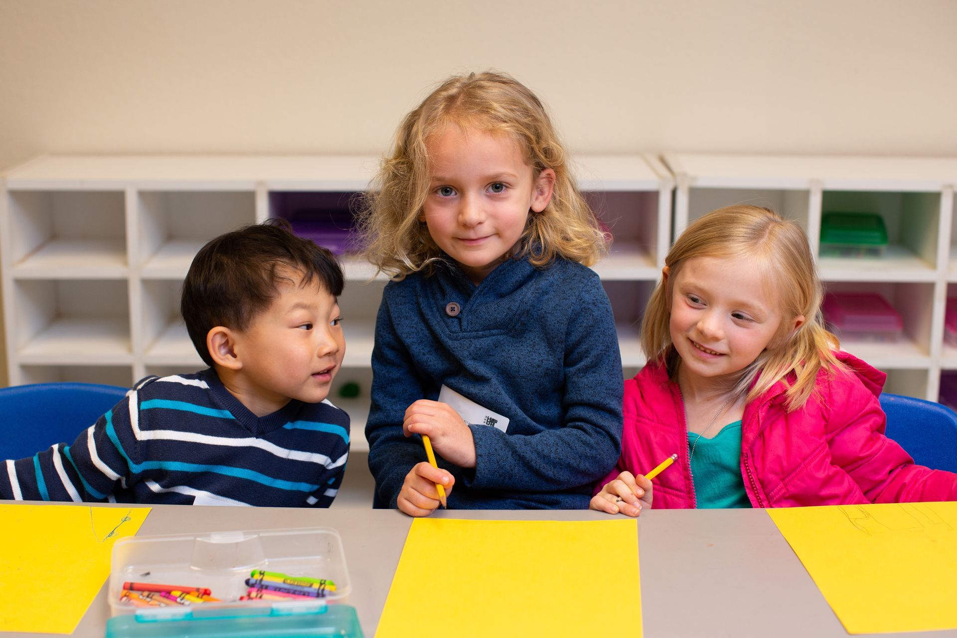 three children are sitting at a table in a classroom holding crayons .