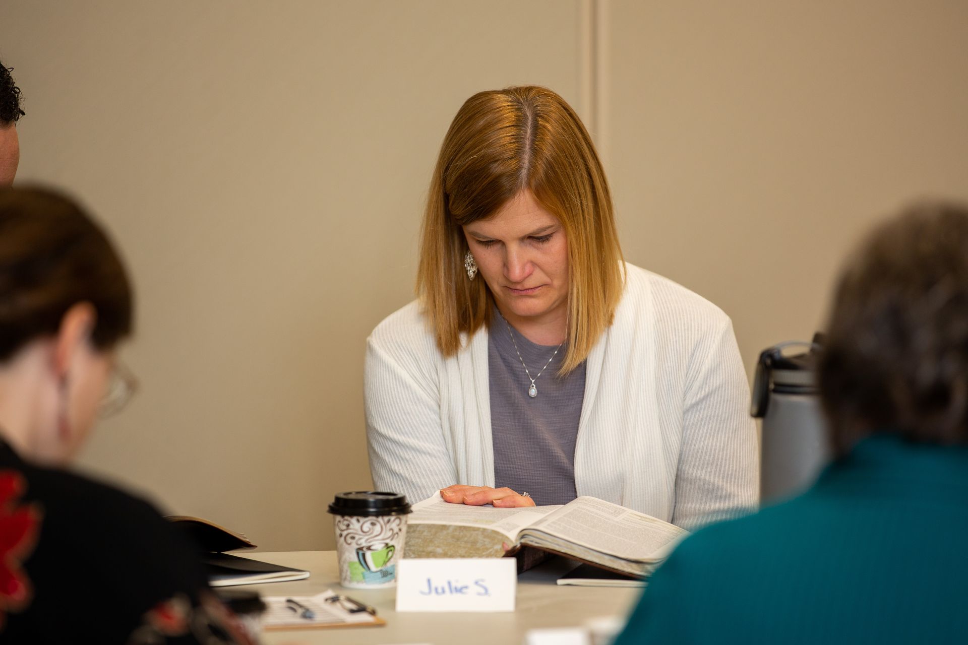 a woman is reading a bible while sitting at a table with other people .