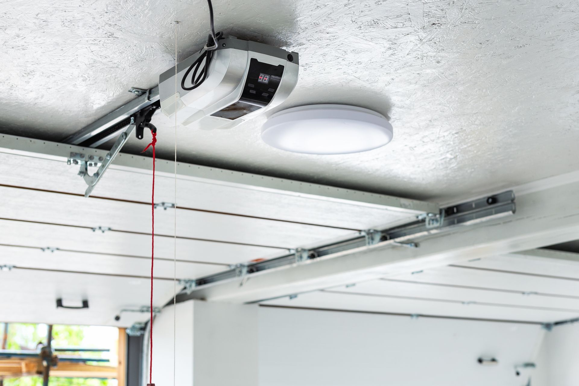 Garage ceiling with overhead door opener and red pull cord, white ceiling light, and metal rails
