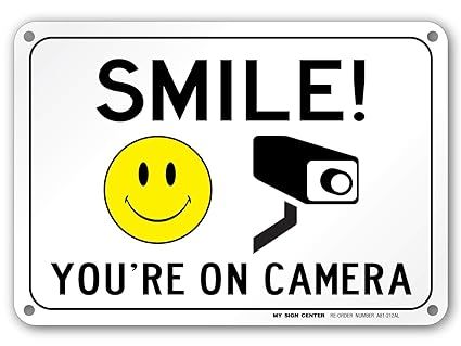 A sign that says `` smile ! you 're on camera '' with a smiley face and a camera.