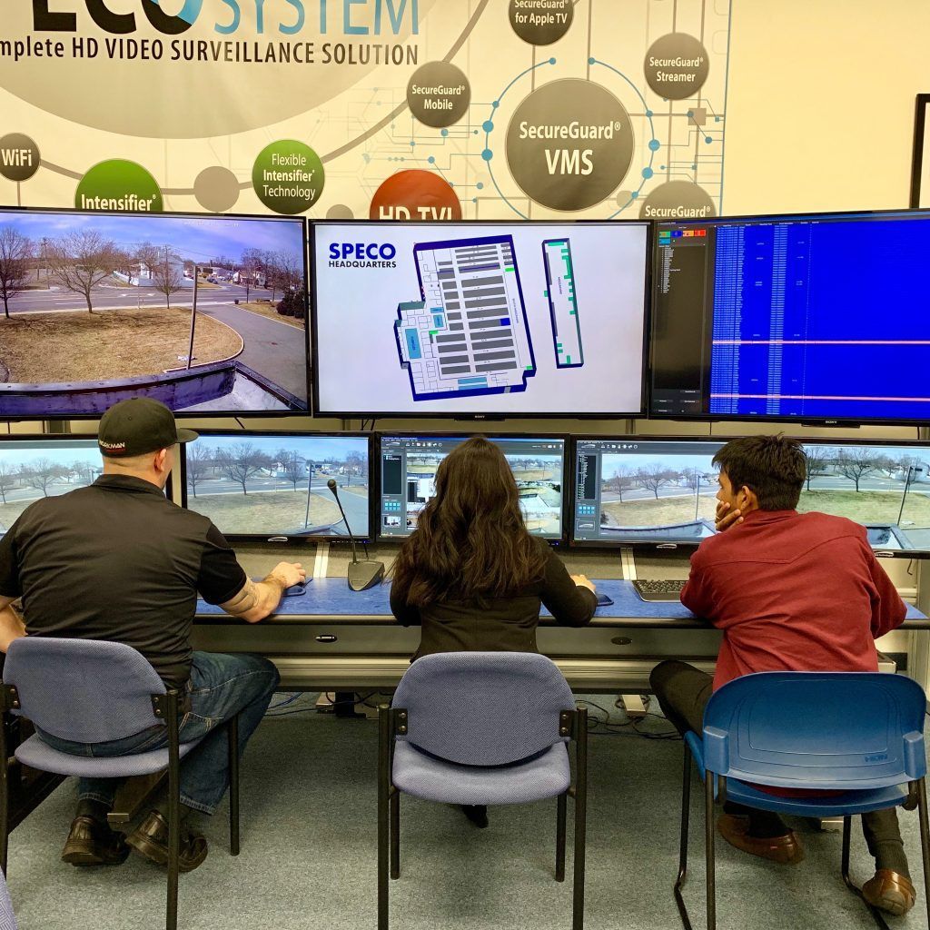 A group of people are sitting at a desk in front of a screen that says speco