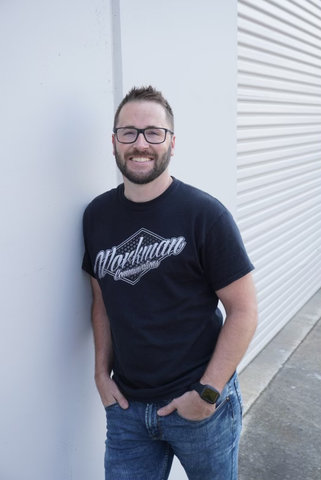 A man wearing glasses and a black shirt is leaning against a wall.