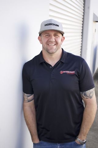 A man wearing a hat and a black polo shirt is standing in front of a white wall.