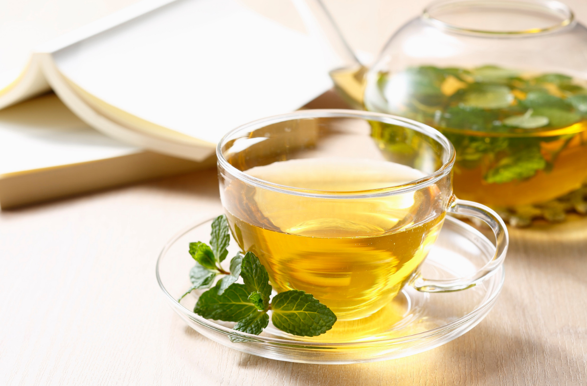 Glass cup and teapot of herbal tea with mint sprig, beside an open book.