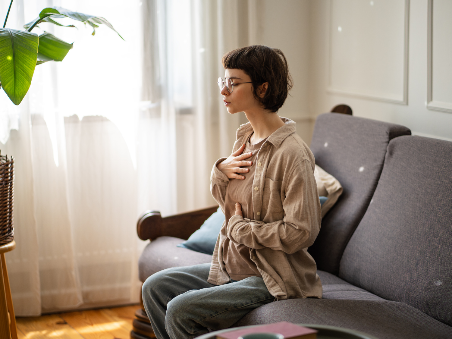 Woman seated on sofa, eyes closed, hands on chest, practicing deep breathing in sunlit room.
