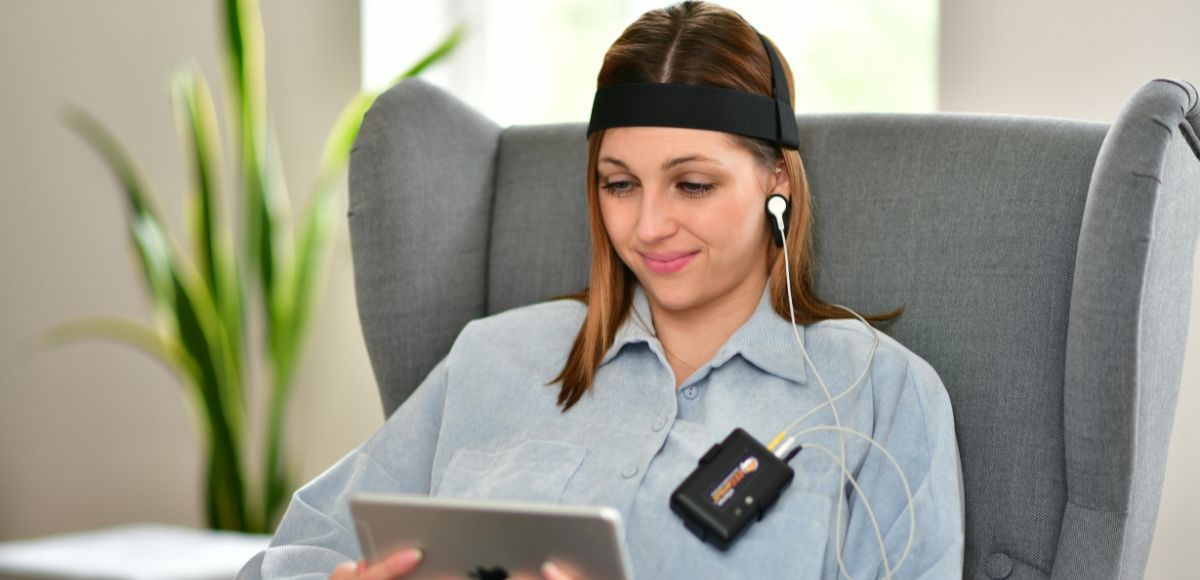 Woman using a tablet while seated in a gray chair, wearing a headset in a bright room.