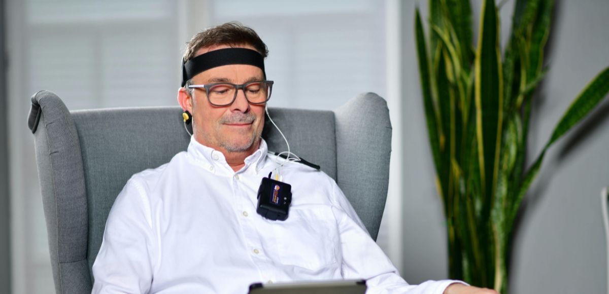 Man seated in a gray armchair, wearing a white shirt and black headband, with a potted plant in the background