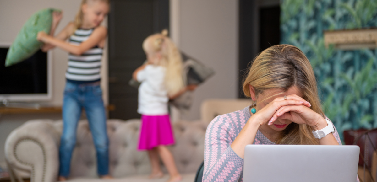 A woman sits at a laptop with her head in her hands while two children have a pillow fight on the couch behind her.