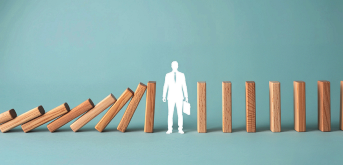Wooden dominoes falling toward a businessperson cutout standing between them on a teal background.