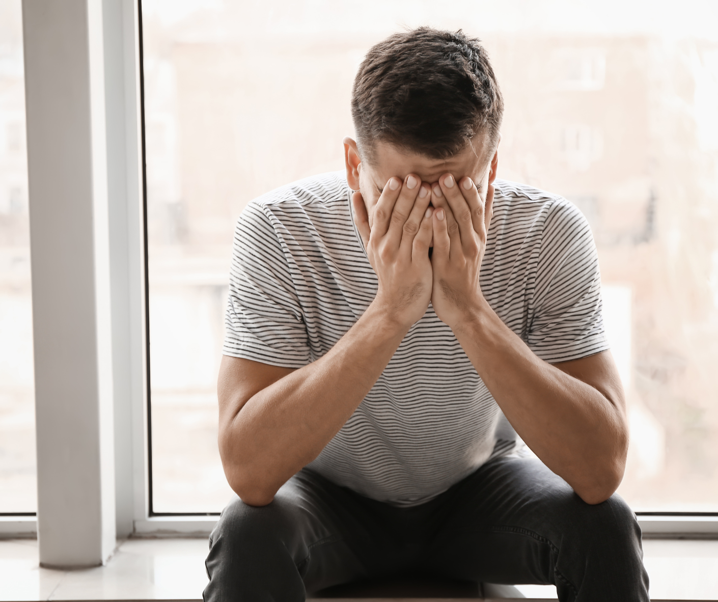 Man sitting with hands covering face near a window, appearing distressed.