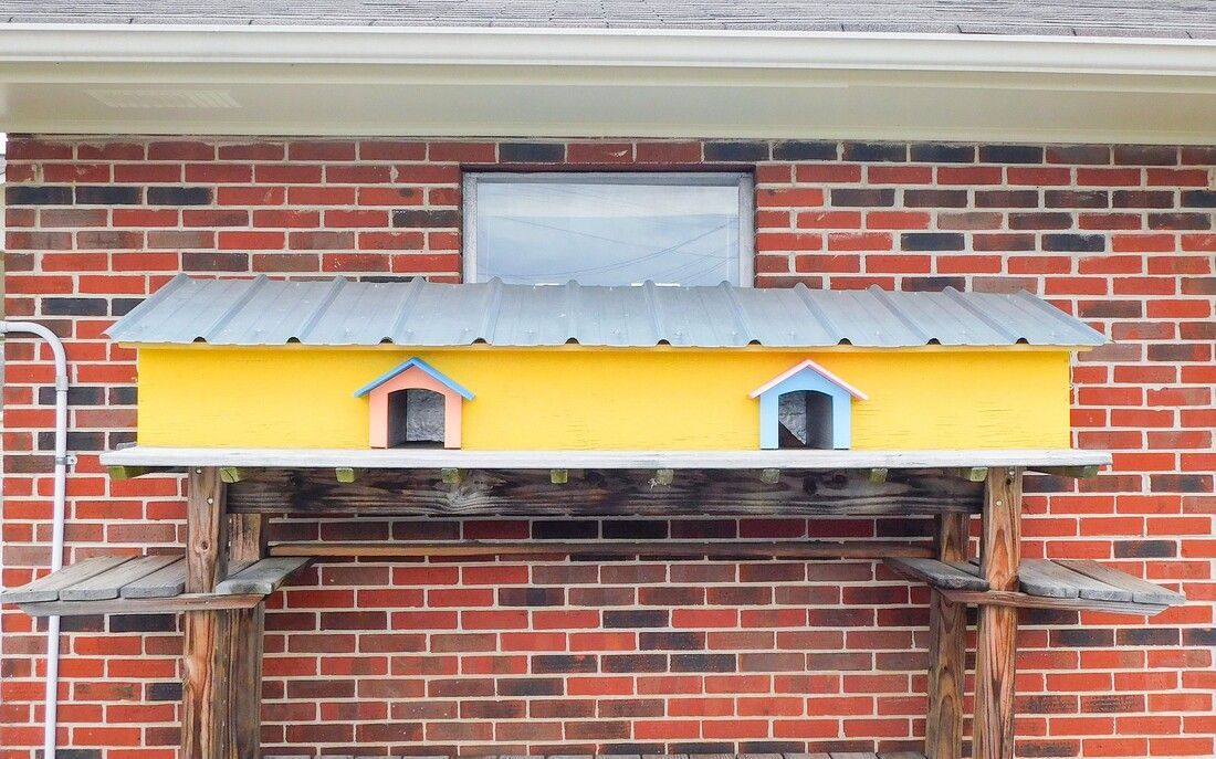 Yellow birdhouse with two windows, attached to a brick wall by a wooden shelf.