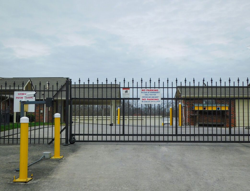 Black metal gate with security spikes, yellow bollards, and buildings under cloudy sky.