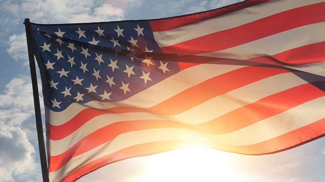 The American flag waves against a bright, sunlit sky with soft clouds in the background.