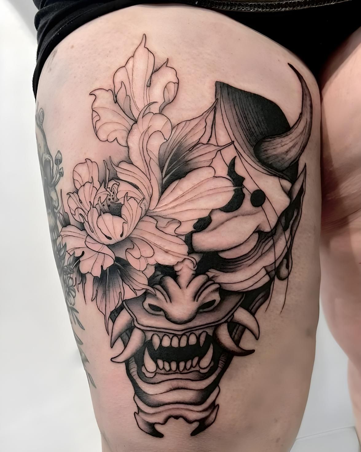 Tattoo of a Japanese oni mask with flowers on a thigh. Black and gray shading with detailed linework — Tattoo Inc Studio In Broadbeach, QLD