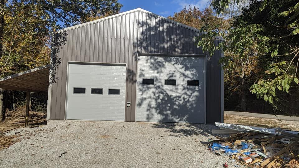 A large metal garage with a white garage door is surrounded by trees.