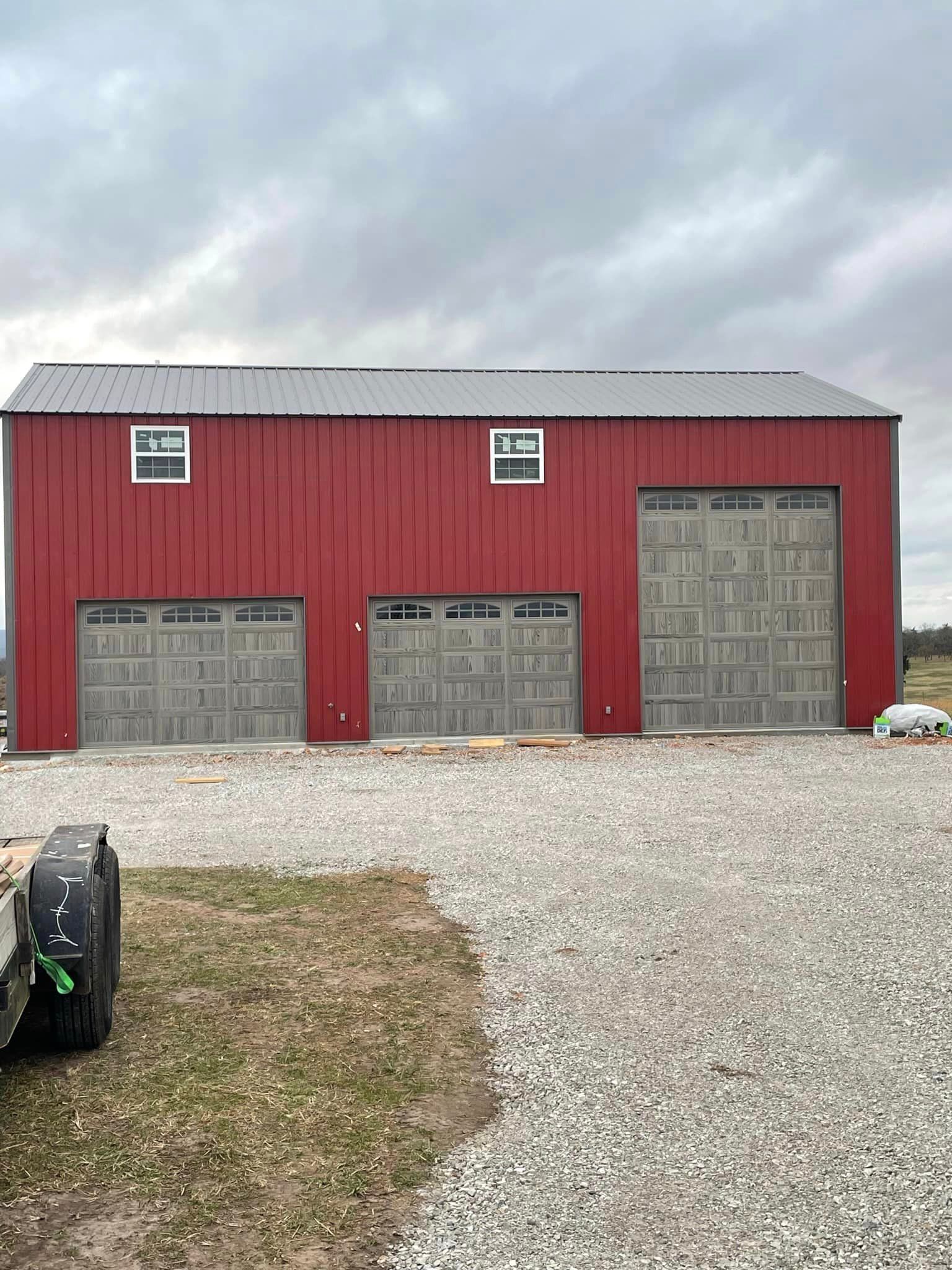Red barn with three gray garage doors on a gravel lot under an overcast sky.