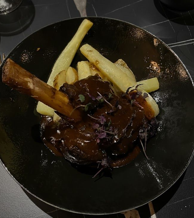 Jarret d'agneau avec sauce et frites, servi sur une assiette noire.