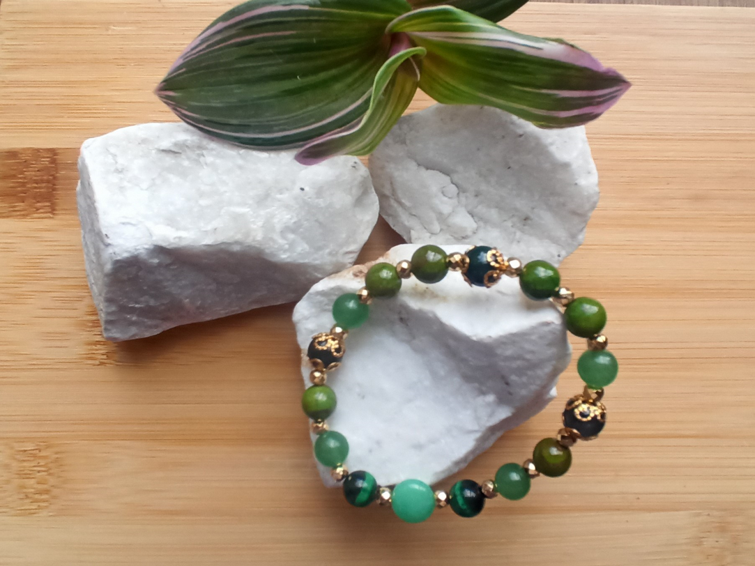 Pulsera de cuentas verdes y doradas sobre piedras blancas con una planta sobre una superficie de madera.