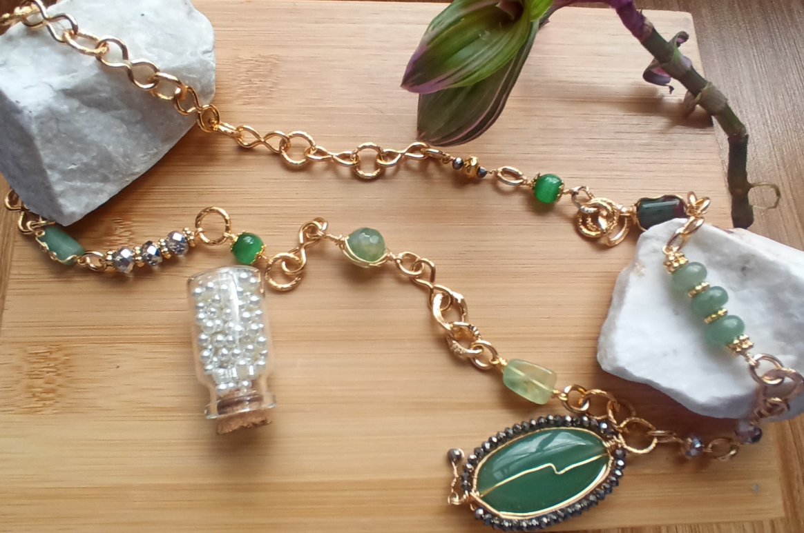 Un collar de cadena de oro con piedras preciosas verdes, una pequeña botella transparente con cristales y un colgante ovalado verde descansa sobre una superficie de madera con piedras blancas.