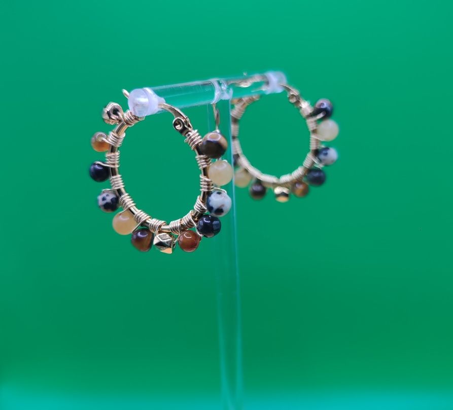 Un par de aros de color dorado con cuentas de colores, exhibidos en un soporte transparente sobre un fondo verde.