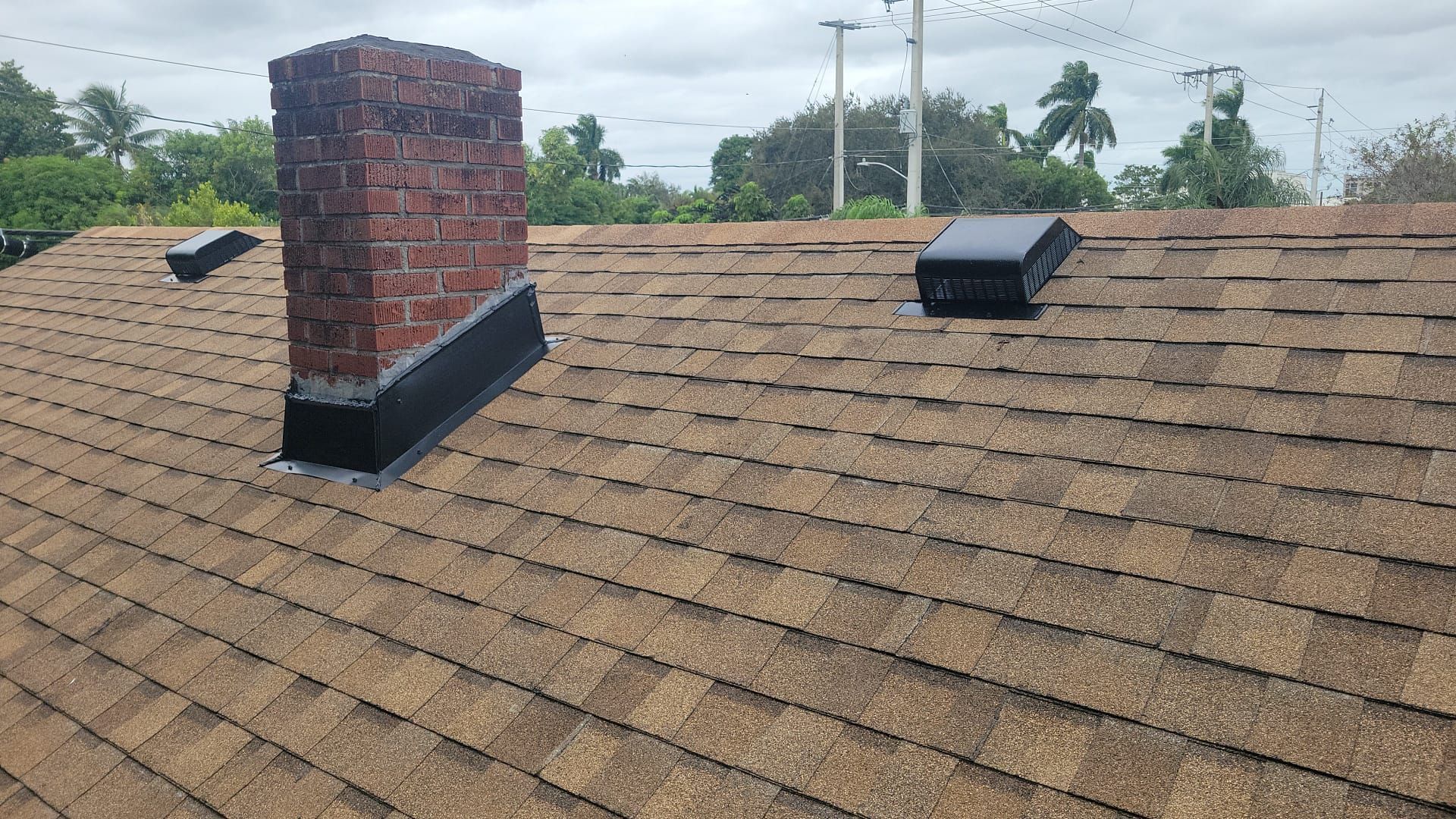 A roof with brown shingles, a brick chimney, and a black vent on a cloudy day.