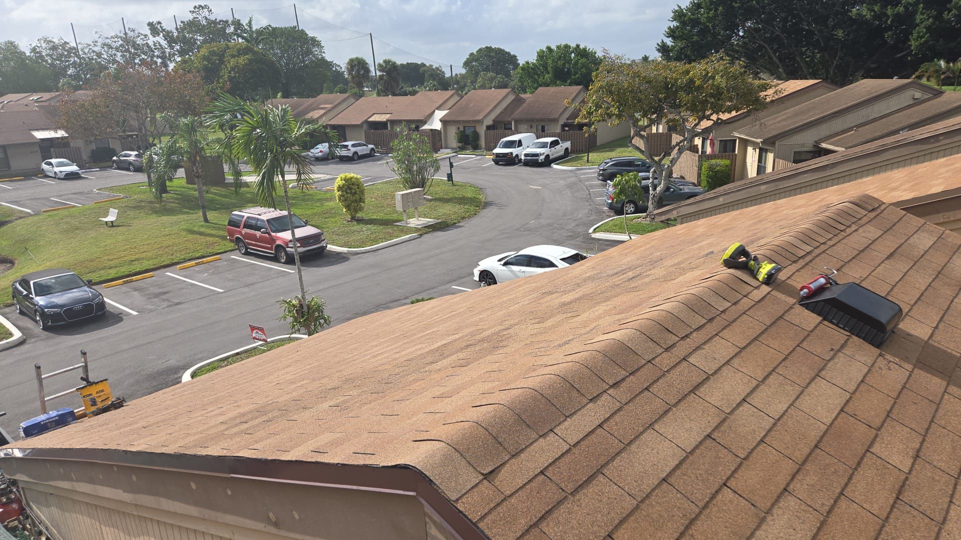 Rooftop view of a residential area with brown houses, asphalt roads, parked cars, and green lawns under a cloudy sky.