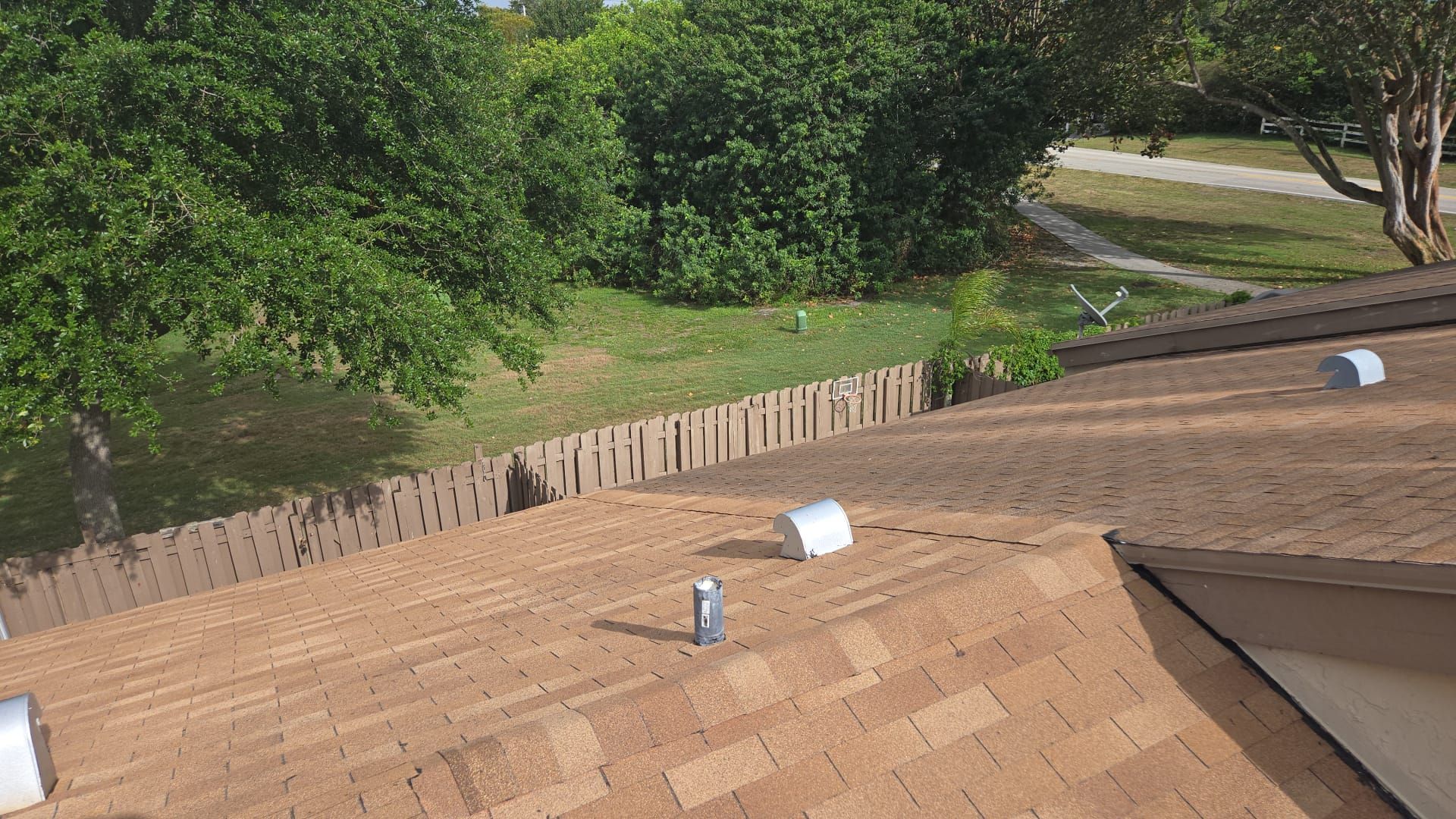 Brown shingle roof with vents, overlooking a backyard with a wooden fence, grass, and trees.
