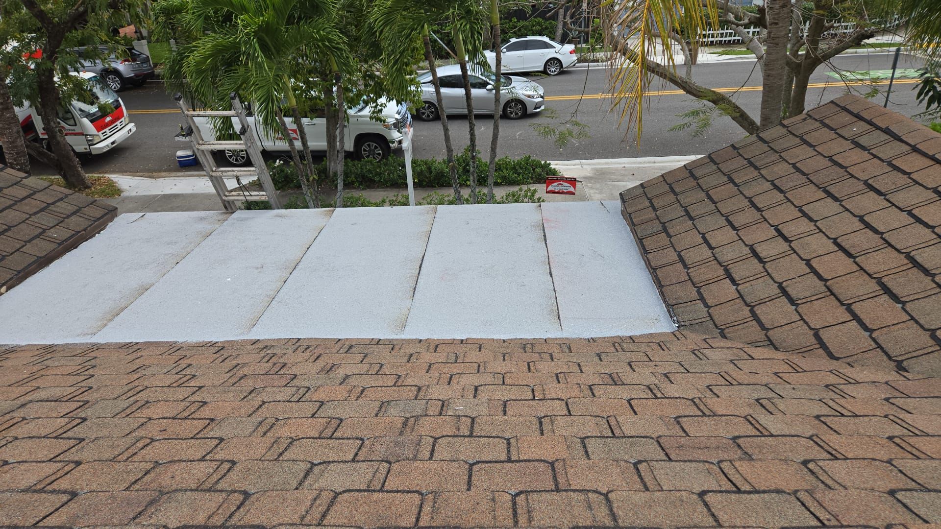 View from a brown shingled roof towards a flat, white roof section with a street and parked cars visible.