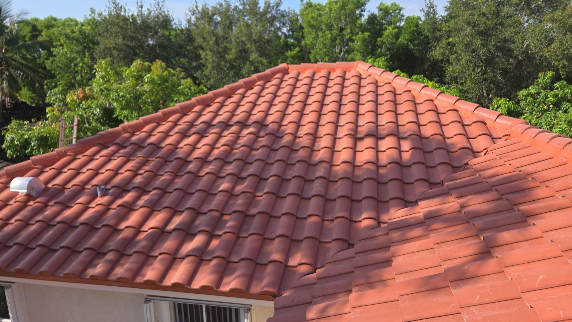 Red tile roof with lush green trees in the background. The roof is slightly angled with sunlight casting shadows.
