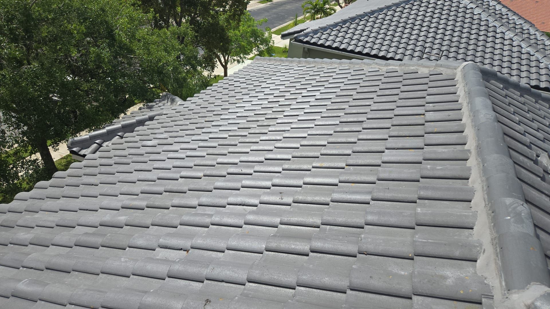 Gray tile roof of a house, with trees and another roof visible in the background on a sunny day.