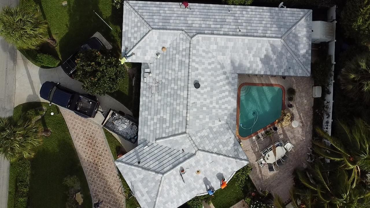Aerial view of a house with workers on the gray roof. There's a pool, trees, and a black truck nearby.