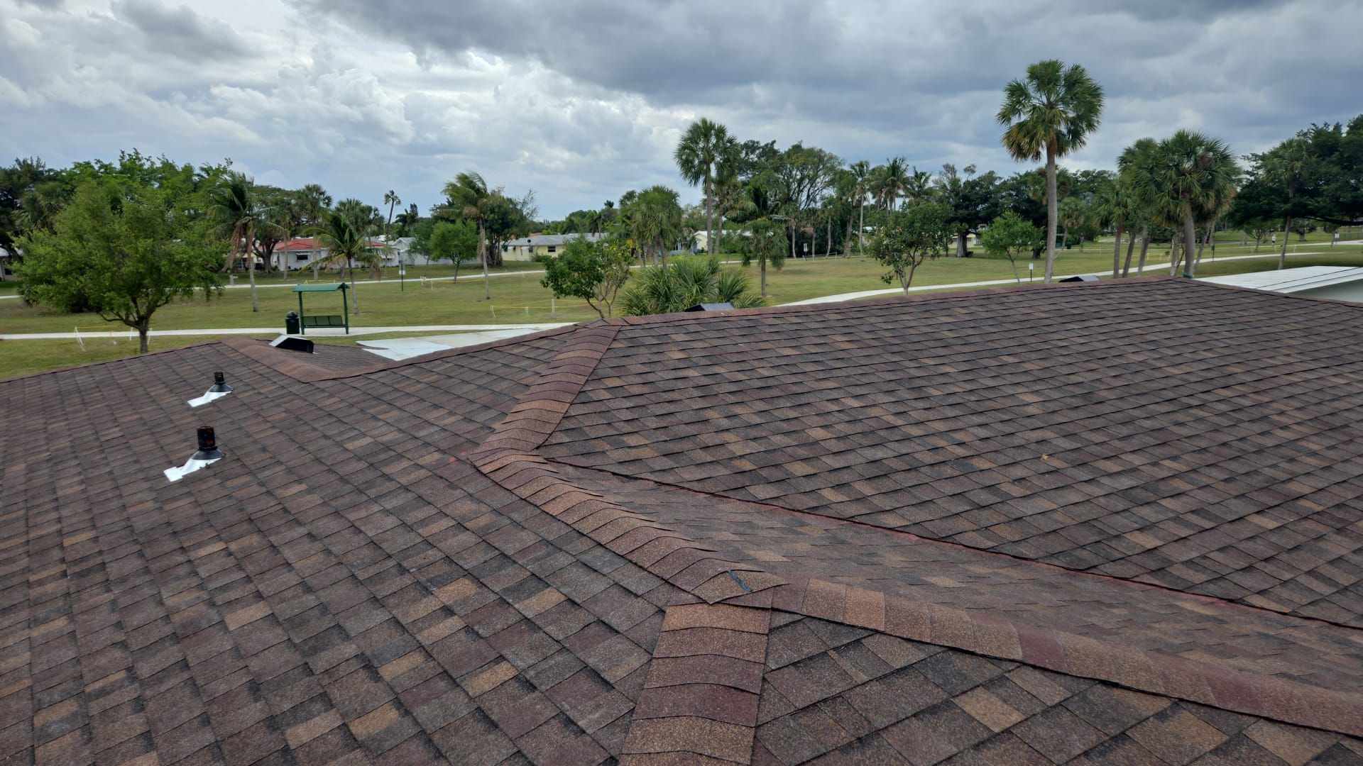A brown shingled roof on a house with a cloudy sky and palm trees in the background.