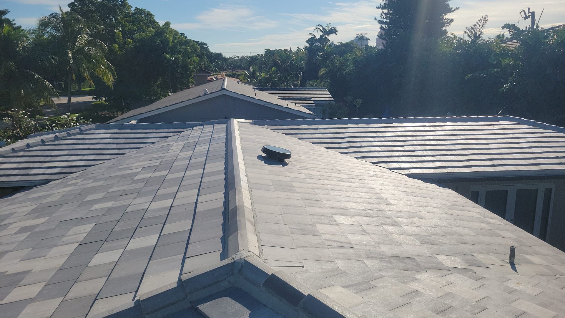 A gray shingled roof with a peak, viewed from above, with trees in the background under a sunny sky.