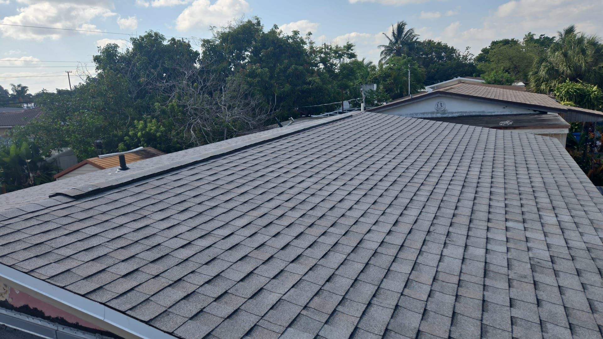 Gray shingle roof with a dark edge in a residential area, trees in the background under a partly cloudy sky.