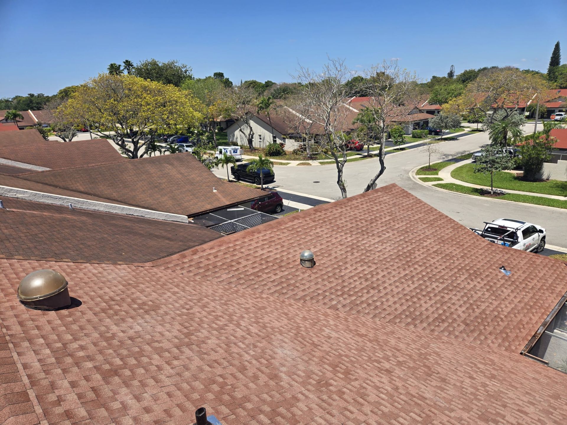 Rooftop view of brown shingle roofs in a suburban neighborhood with trees and a street visible under a blue sky.