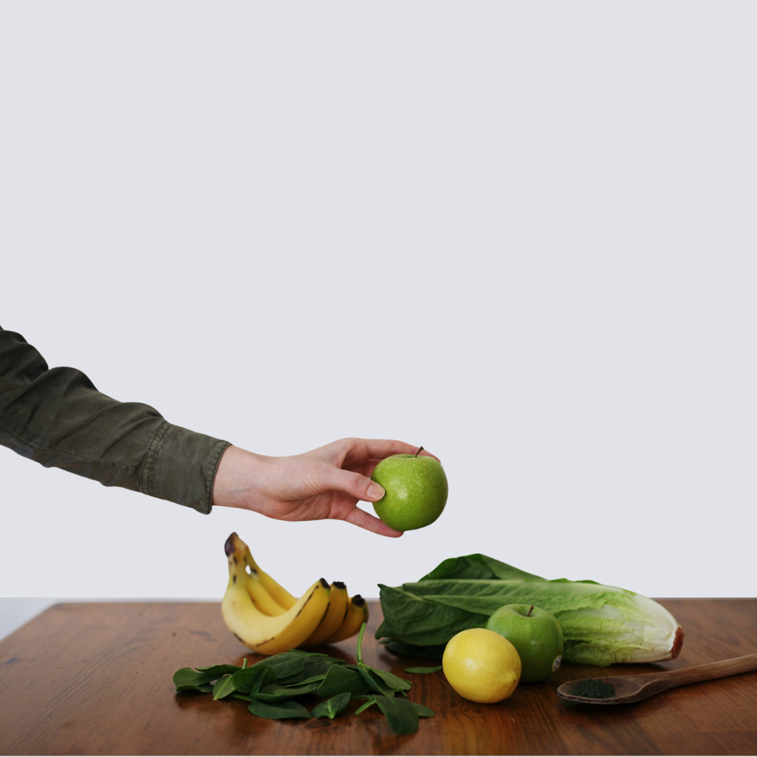 hand holding a green apple above various fruits and vegetables on a table