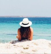 A woman wearing a hat is sitting on the beach looking at the ocean.