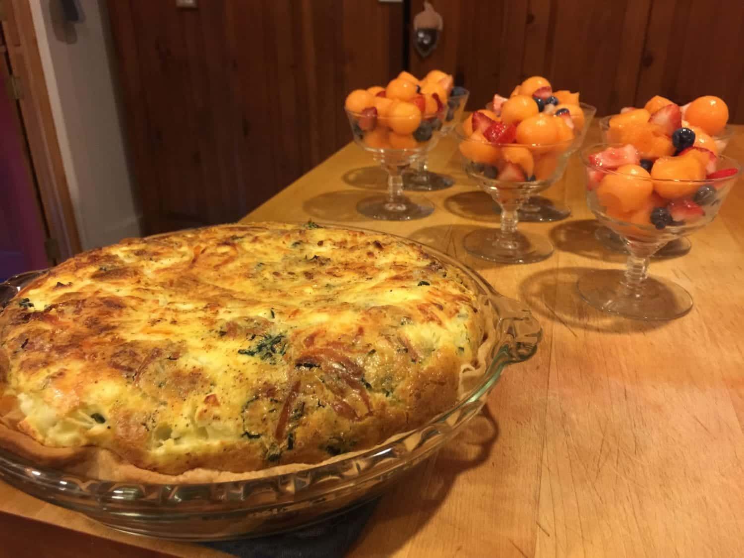 A quiche is sitting on a wooden table next to bowls of fruit.