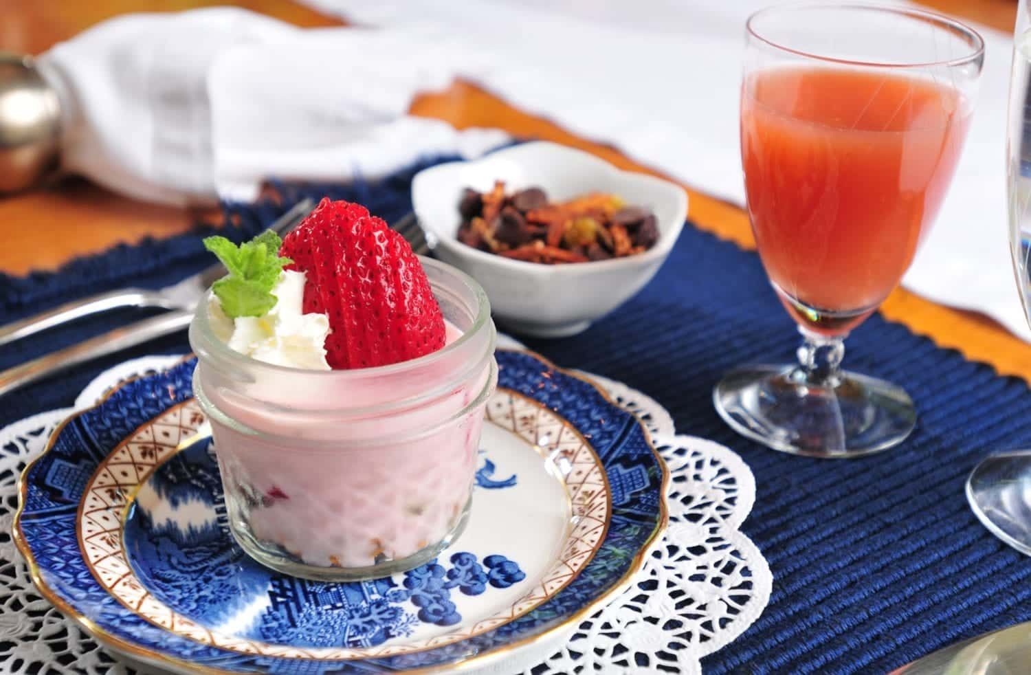 A jar of yogurt with whipped cream and strawberries on a plate next to a glass of juice.