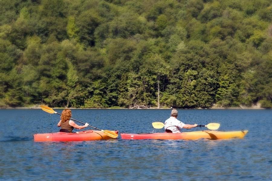Two people are kayaking on a lake with trees in the background.