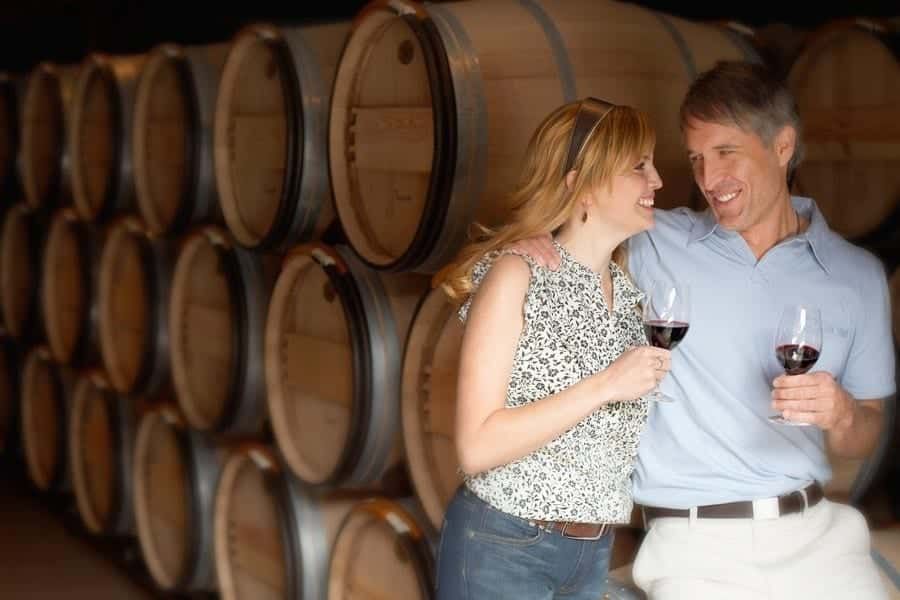 A man and a woman are holding wine glasses in front of a stack of wine barrels.