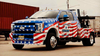 Black and white tow truck with American flag design parked on snow-covered ground under a cloudy sky.