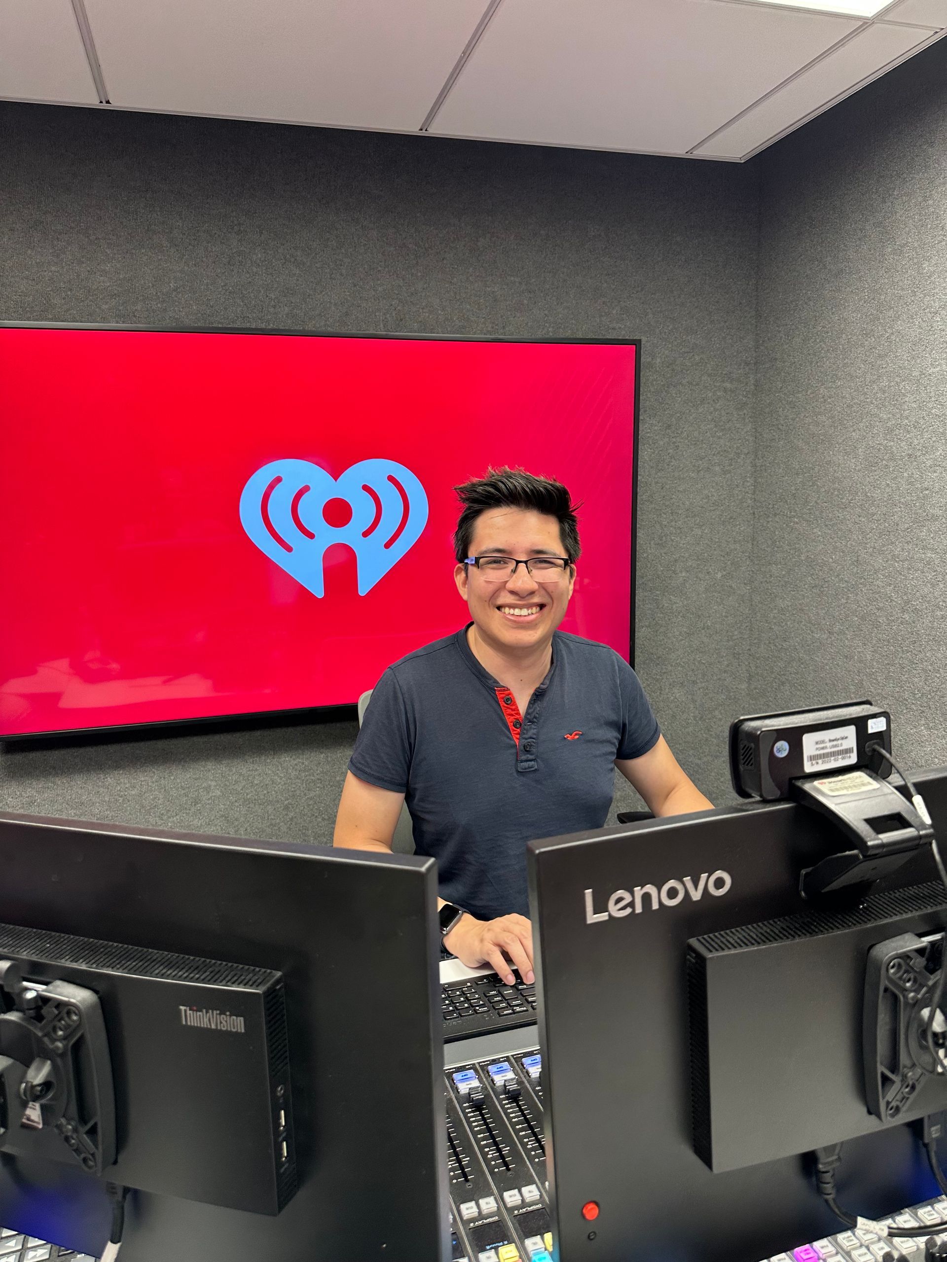 Person smiling behind a soundboard in a studio, with a red screen showing a white heart logo.